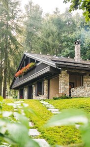 Uno chalet circondato dalla foresta. Le pareti esterne sono rivestite in pietra, i balconi e il tetto sono in legno. | © 33COM srls