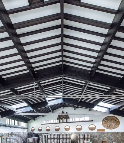 Die Dachstruktur aus Holz einer großen Produktionshalle mit dunkel lackierten Leimbindern, Pfetten und Sparren | © Benjamin Pfitscher