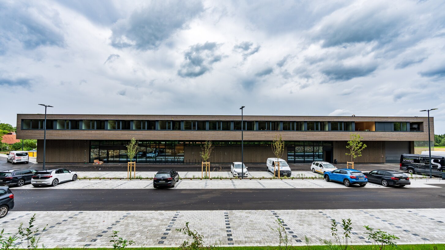 Ein breites zweistöckiges Gebäude mit einer Fassadenverkleidung aus Holzleisten, davor ein Parkplatz | © Roland Wehinger