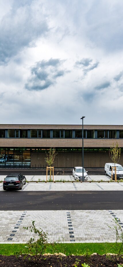 Un ampio edificio a due piani con un rivestimento di facciata in doghe di legno e un parcheggio antistante | © Roland Wehinger