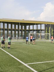 Ein Fußballfeld mit Spielern, im Hintergrund ein großes Gebäude mit vielen Fenstern und einem goldig glänzenden Dach | © Michele Nastasi