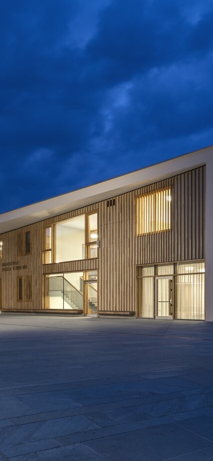 A large building with a white painted exterior wall and wooden cladding on the façade. Light burns inside | © Oliver Jaist