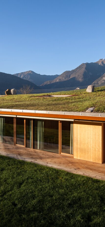 Ein Gebäude, das in den Hang gebaut ist. Sichtbar sind nur eine Fensterfront mit Holzfenstern, das Dach ist begrünt. Berge im Hintergrund. | © Davide Cornacchini